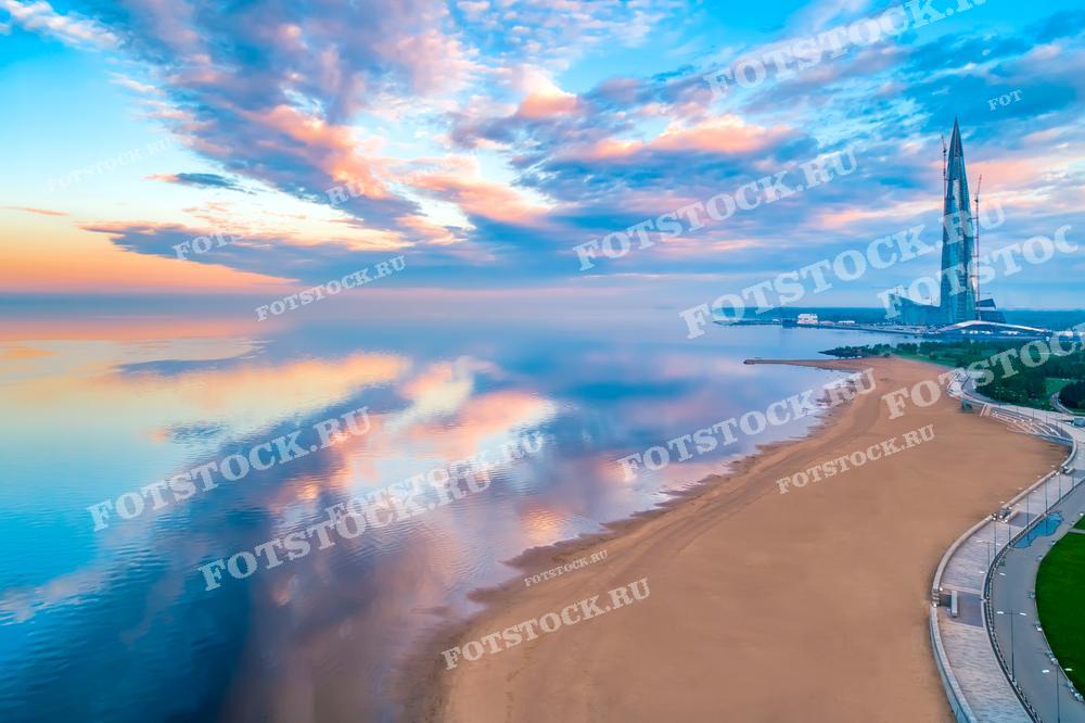 На фотографии изображена рассветная или закатная сцена на побережье. Высокий небоскрёб Лахта Центра возвышается на фоне красивого неба с разноцветными облаками, отражающимися в спокойной водной глади. Пустынный пляж и променад создают ощущение тишины и умиротворения. Такой снимок может быть использован в качестве рекламы для туристических агентств, в путеводителях, как элемент дизайна интерьера или как вдохновляющий образ в социальных сетях.